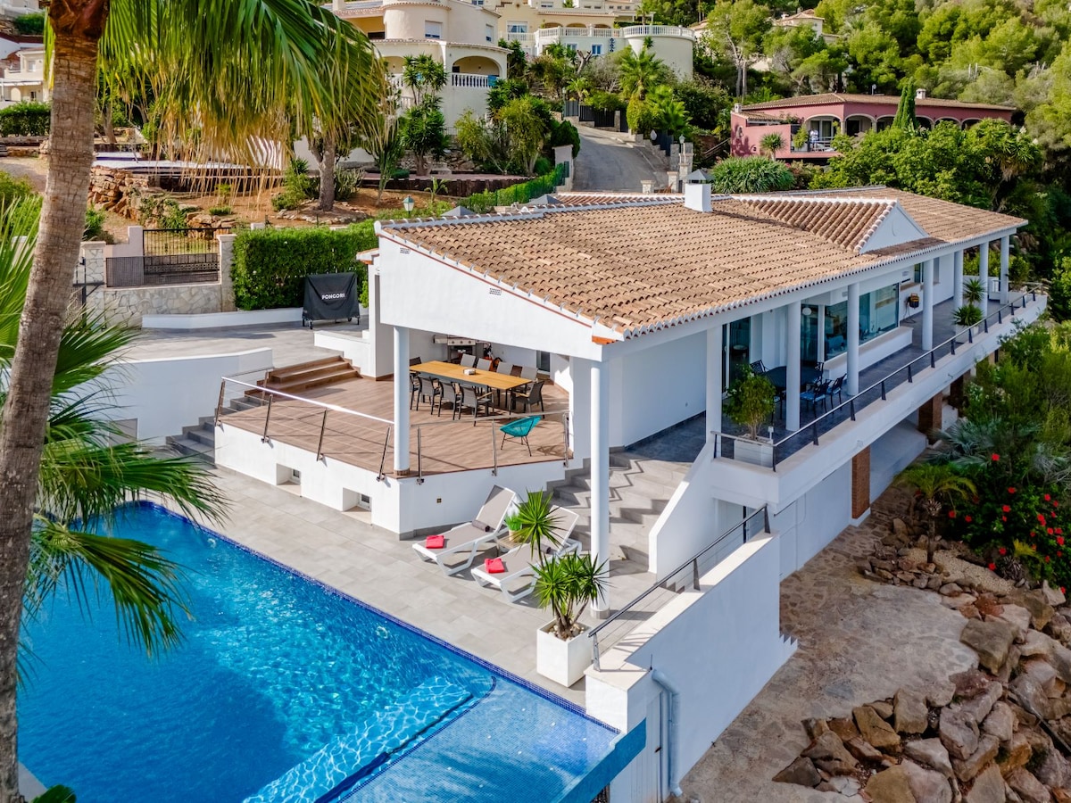 An aerial view captures a modern villa featuring a private pool and expansive outdoor deck. Lush greenery surrounds the property, while the inviting pool reflects sunlight. A large dining table is visible on the deck, offering ample space for outdoor gatherings.