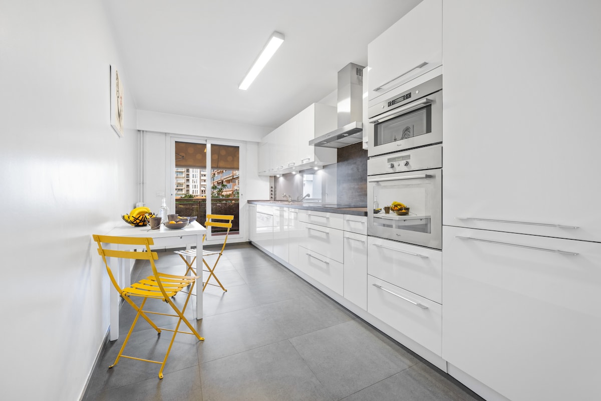 An open kitchen is displayed, featuring modern white cabinetry and stainless steel appliances. Two yellow folding chairs are arranged around a small table, with a bowl of fruit visible. Large windows provide natural light and views of the outside.