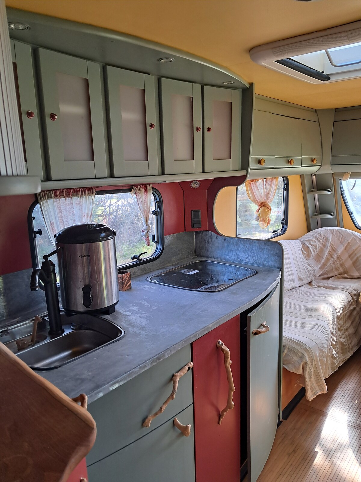 The interior of the caravan features a compact kitchen area with a sink, stove, and a kettle. Light green cabinetry offers ample storage, complemented by a cozy seating area with soft cushions. Natural light enters through the window, enhancing the functionality of the space.