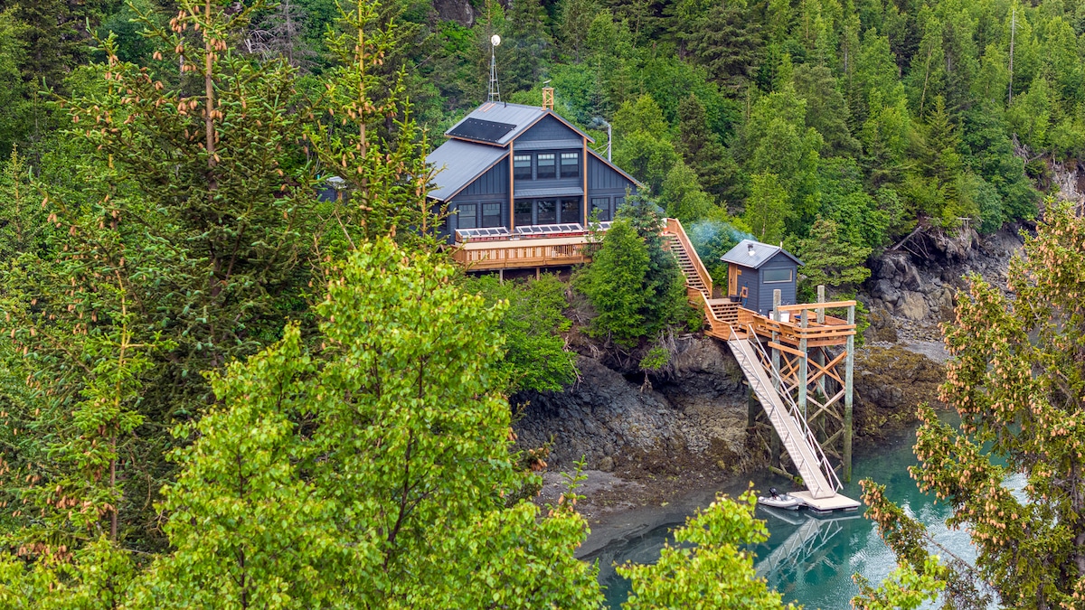 The property is nestled among lush greenery, featuring a modern home with expansive windows. A wooden deck extends from the house, leading to a multi-step staircase that descends to a rocky shore by the water's edge.