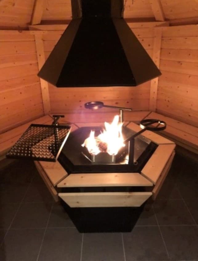 An inviting fire pit is centrally located within a wooden structure, surrounded by a hexagonal seating area. The fire is lit, providing warmth and a focal point. Wooden walls create a cozy atmosphere, while a ventilation hood ensures airflow, enhancing the experience.