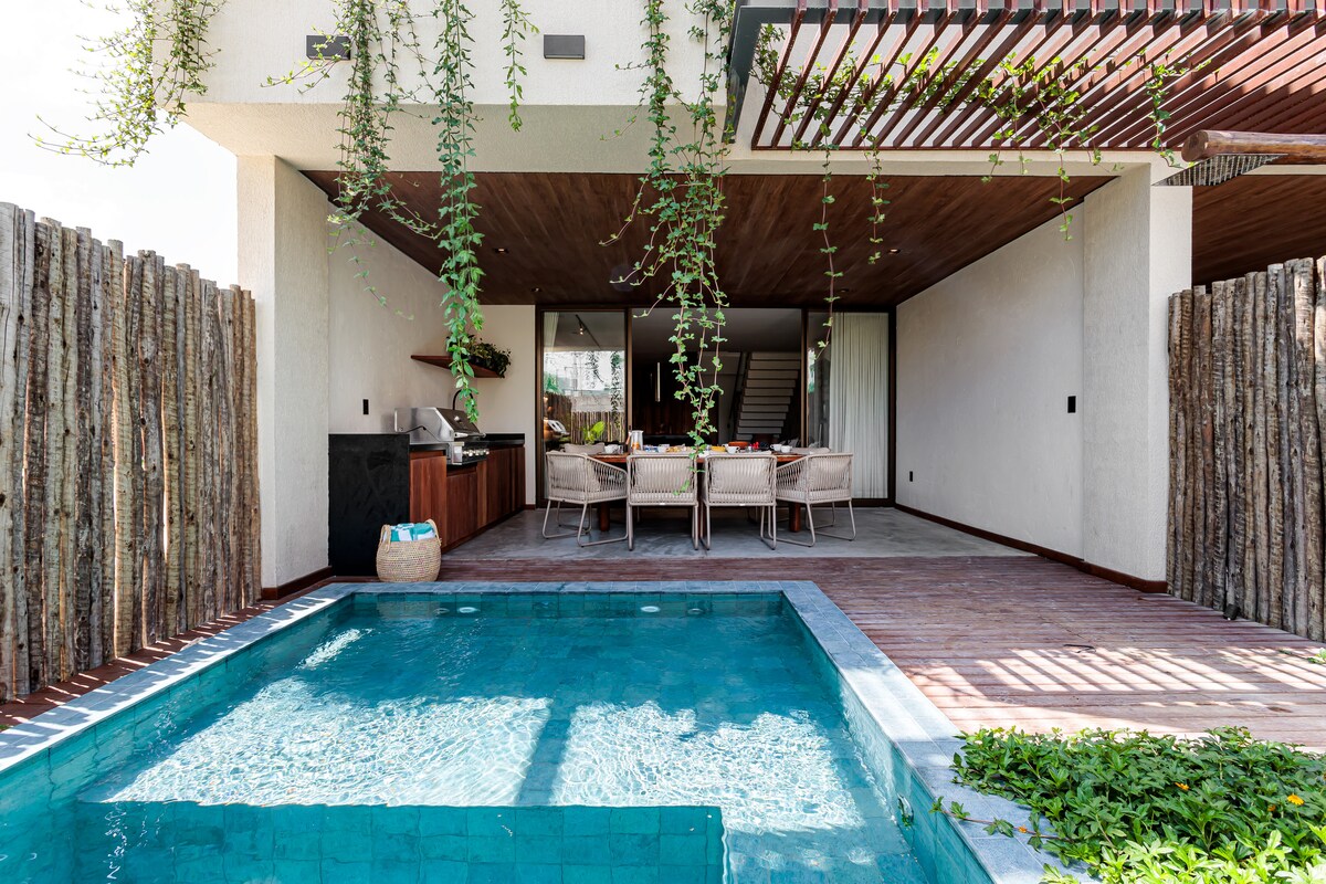 An outdoor area with a small turquoise pool is complemented by a dining table set for multiple guests. Wooden beams and a natural fence provide privacy, while greenery hangs from the ceiling, enhancing the space's relaxed ambiance.