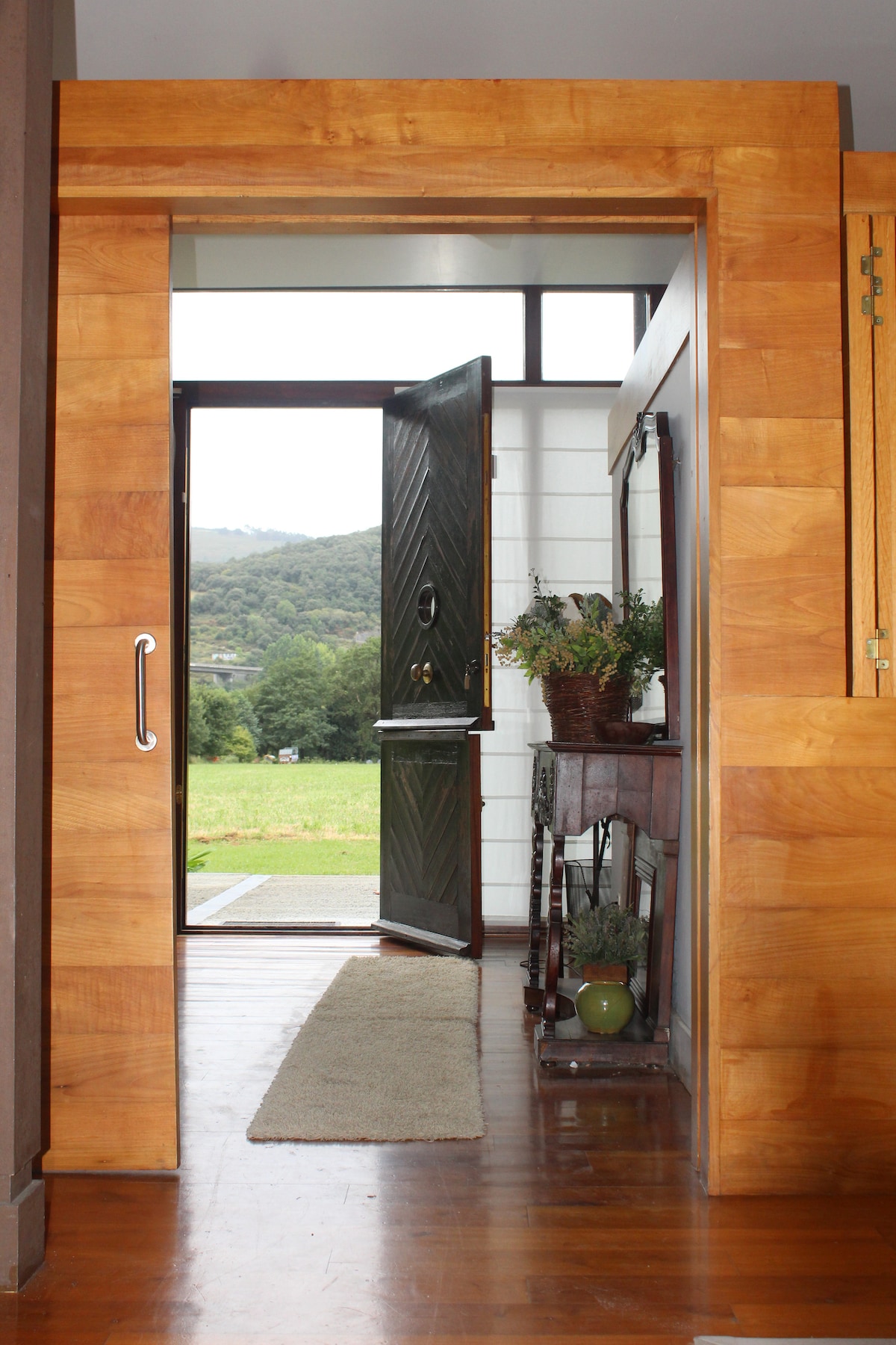 A wooden door with a diagonal design opens into a welcoming entryway. Natural light filters through large windows, illuminating the polished floor. A small console table, adorned with a decorative plant, sits to the side, while a soft area rug lies in front of the door.
