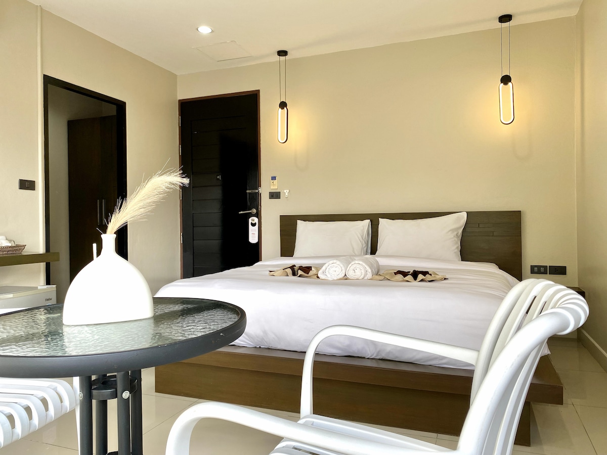 A bright sleeping area features a spacious bed adorned with white linens and decorative towels. Minimalist lighting fixtures and a small table with white chairs enhance the clean design. Natural light fills the room from a window, complementing the neutral color palette.