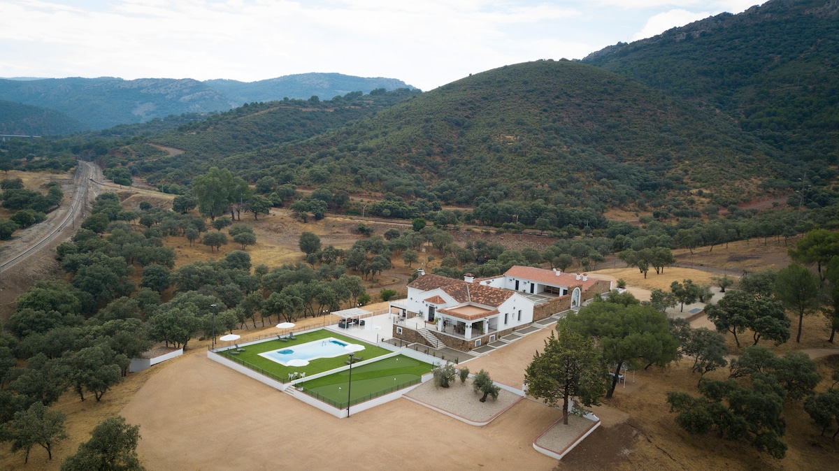 An aerial view captures a spacious property surrounded by rolling hills and trees. The layout includes a large swimming pool, multiple buildings, and well-maintained outdoor space, with natural landscape providing a serene backdrop.