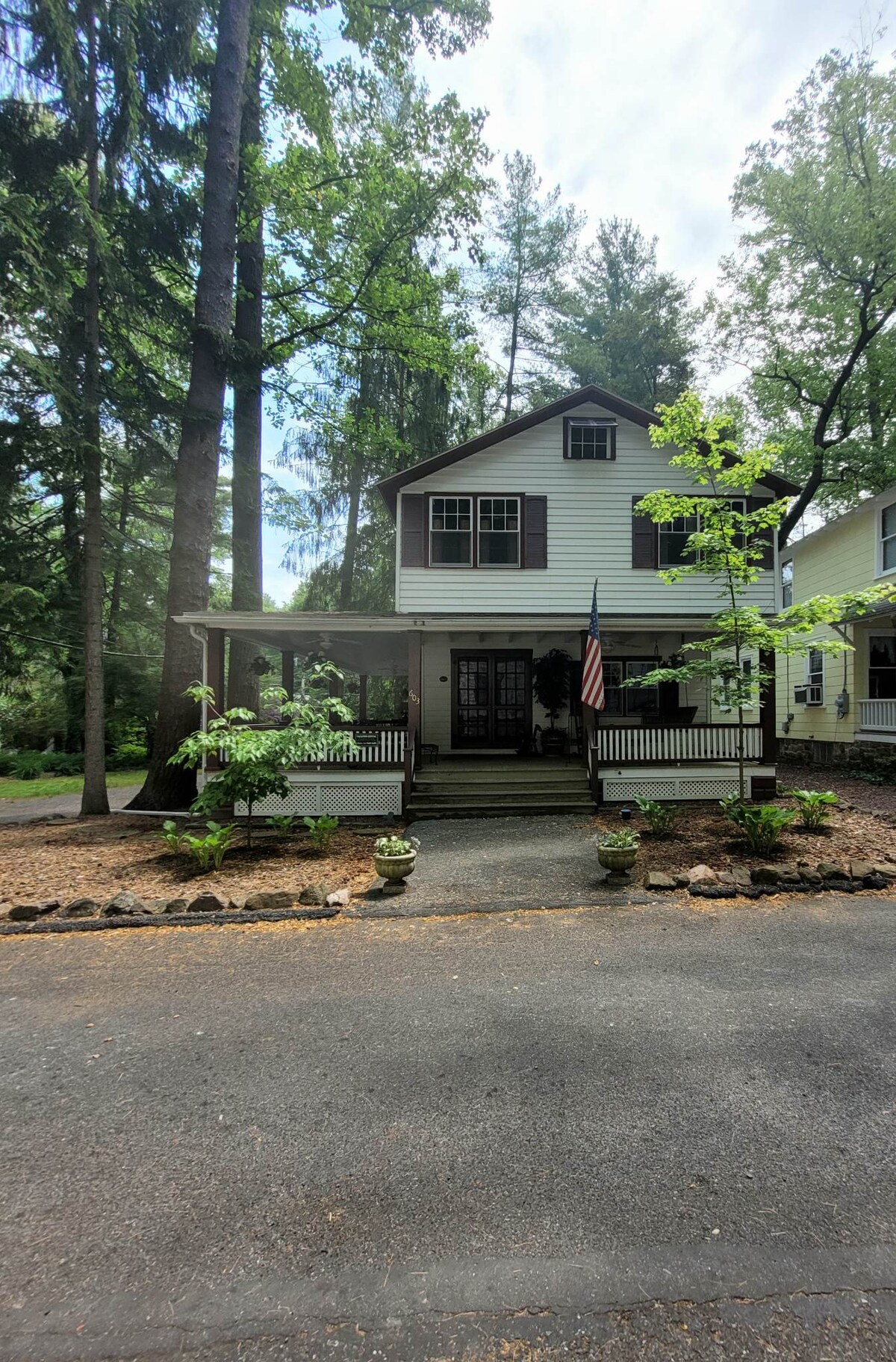 The exterior of the cottage features a two-story design with a welcoming porch adorned with potted plants. A flag is displayed, and tall trees provide natural shade, enhancing the peaceful setting. The pathway leads to the entrance, inviting guests to explore further.