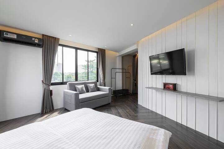 A modern bedroom features large windows that allow natural light to brighten the space. A spacious bed is positioned beside a flat-screen TV mounted on the wall, while a comfortable sofa sits in the corner under soft drapes. Wood flooring adds warmth to the area.
