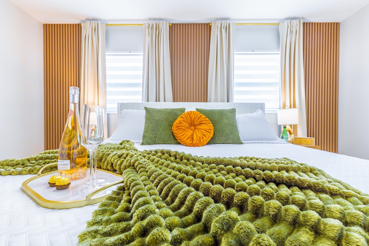 A well-appointed bedroom is showcased with a spacious bed adorned with a textured green throw and a vibrant orange pillow. Two windows with sheer curtains provide natural light, while bedside lamps add a soft glow. A tray with champagne and glasses sits invitingly atop the bed.