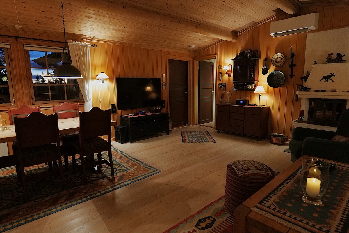 A cozy living area features wooden paneling and warm lighting. A dining table with four chairs is positioned beside a television. Decorative items and a fireplace contribute to the rustic ambiance. Large windows invite natural light and offer views of the outside.