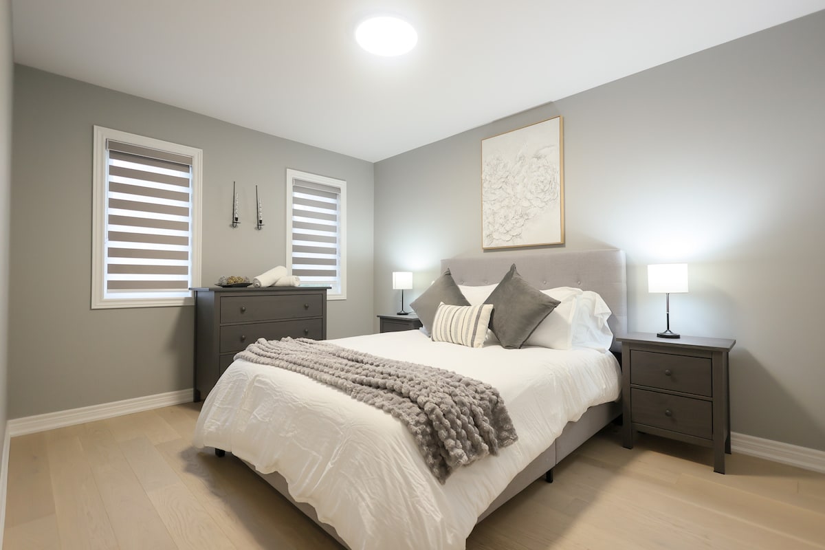 Habitación acogedora con una cama lujosa con una manta acogedora, almohadas decorativas, mesitas de noche individuales con lámparas, un tocador elegante, arte en la pared y persianas modernas.