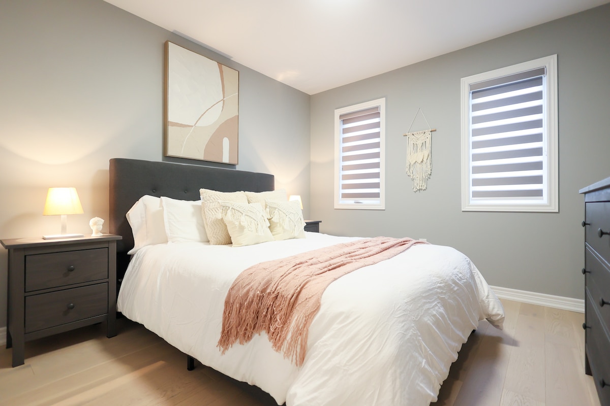 Habitación acogedora con una cama lujosa, almohadas y mantas de textura suave, dos mesitas de noche a juego con lámparas, arte abstracto en la pared, ventanas luminosas con persianas y detalles decorativos acogedores.