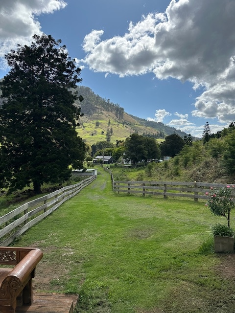 A scenic view of the property showcases lush green lawns bordered by wooden fencing. Rolling hills and trees create a natural backdrop under a partly cloudy sky. The pathway leads towards distant structures, inviting exploration of the expansive landscape.