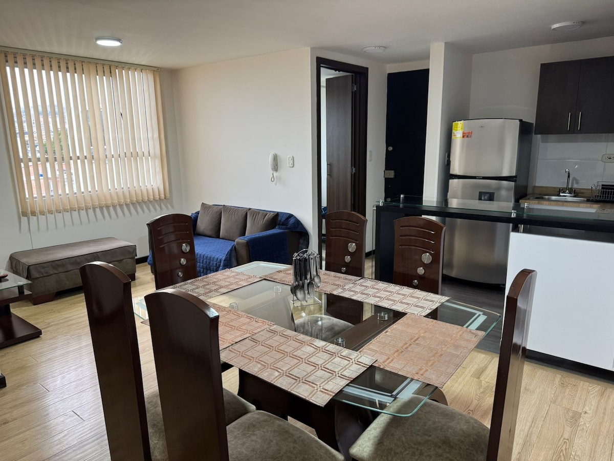 The open living and dining area features a glass dining table surrounded by eight wooden chairs. A blue sofa is positioned near a large window with vertical blinds. An adjacent kitchen area is equipped with modern appliances, including a refrigerator.