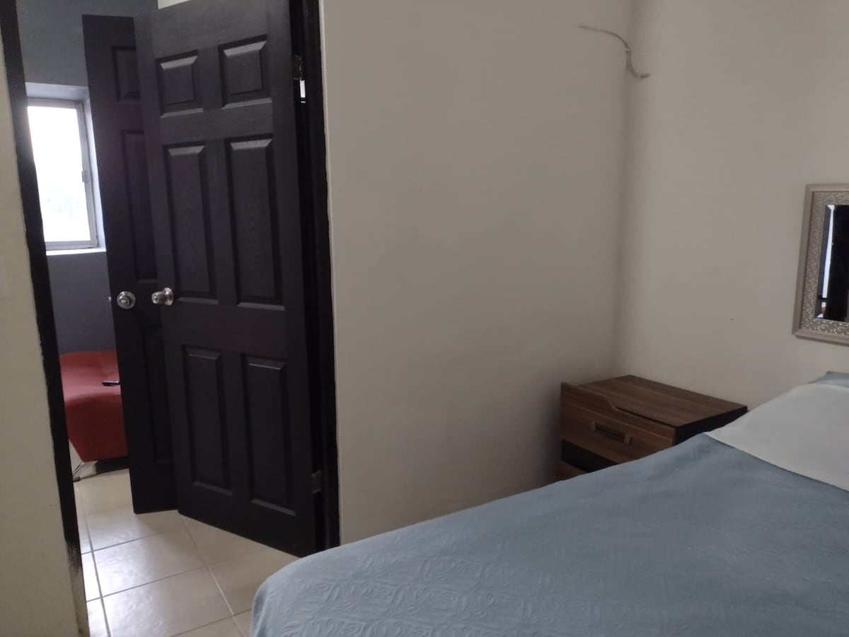 A cozy sleeping area is visible, featuring a light blue bedspread over a spacious bed. A wooden nightstand with drawers is positioned beside the bed. A partially open black door leads to another room, and a small window allows natural light to enter.