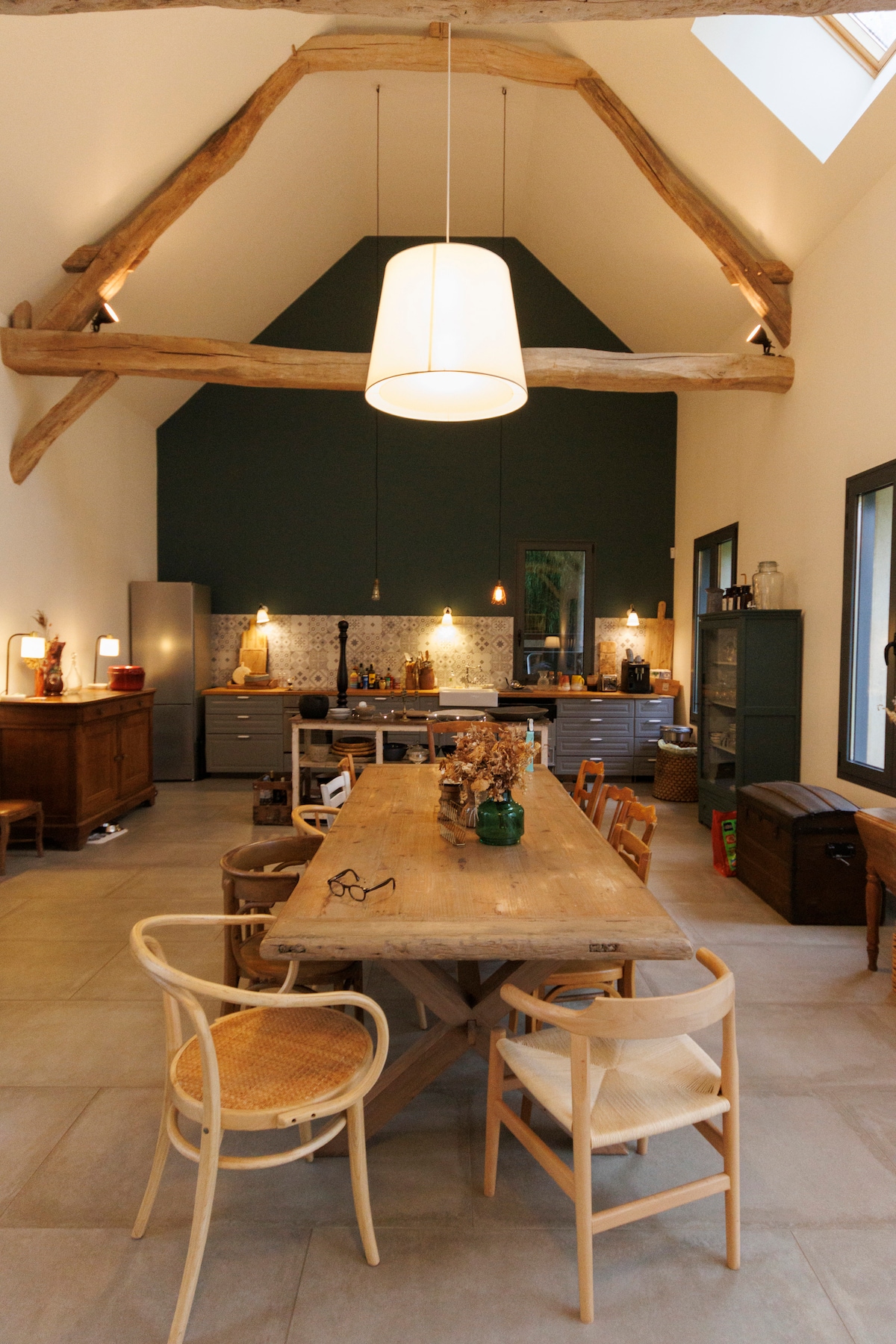A spacious dining area features a long wooden table surrounded by chairs. A pendant light hangs above, while large windows allow natural light to flood in. The high, wooden-beamed ceiling enhances the room's openness, and a well-equipped kitchen is visible in the background.