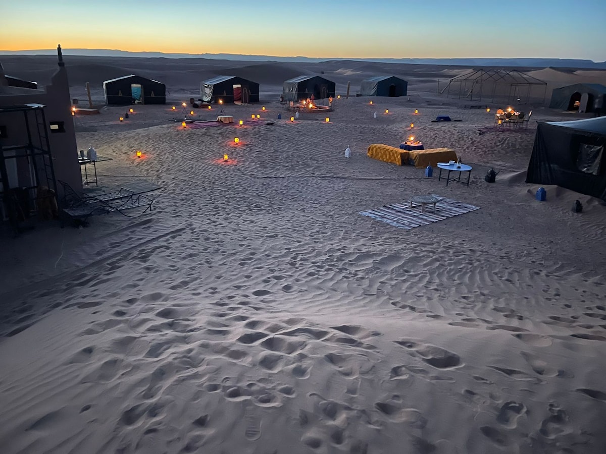 A tranquil desert setting at dusk showcases several tents arranged amidst gently rolling sand dunes. Softly flickering candlelight illuminates the area, creating a serene atmosphere. Footprints in the sand lead through the expanse, emphasizing the quiet and peaceful nature of the landscape.