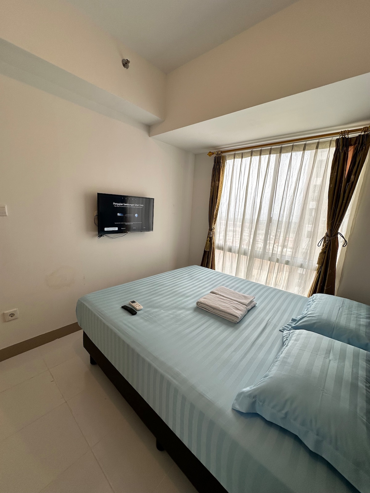 A cozy bedroom features a spacious bed with light blue linens and neatly folded towels. Large windows, adorned with curtains, allow natural light to fill the room. A wall-mounted television is positioned opposite the bed, enhancing the comfortable atmosphere.