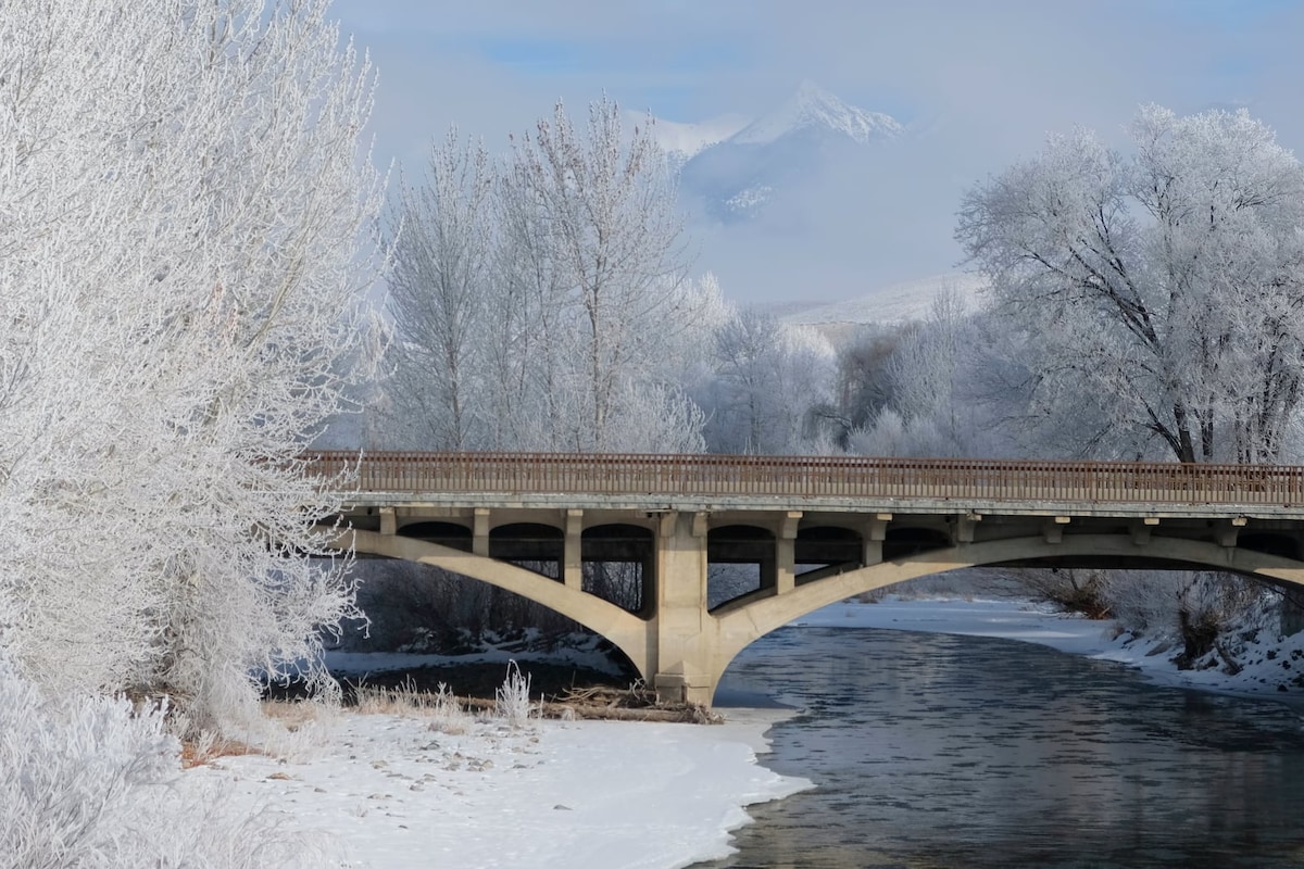 A bridge spans across a tranquil river, framed by frosted trees and distant mountains. The soft white landscape is enhanced by the crispness of the winter scene, revealing a still waterway reflecting the serene surroundings.