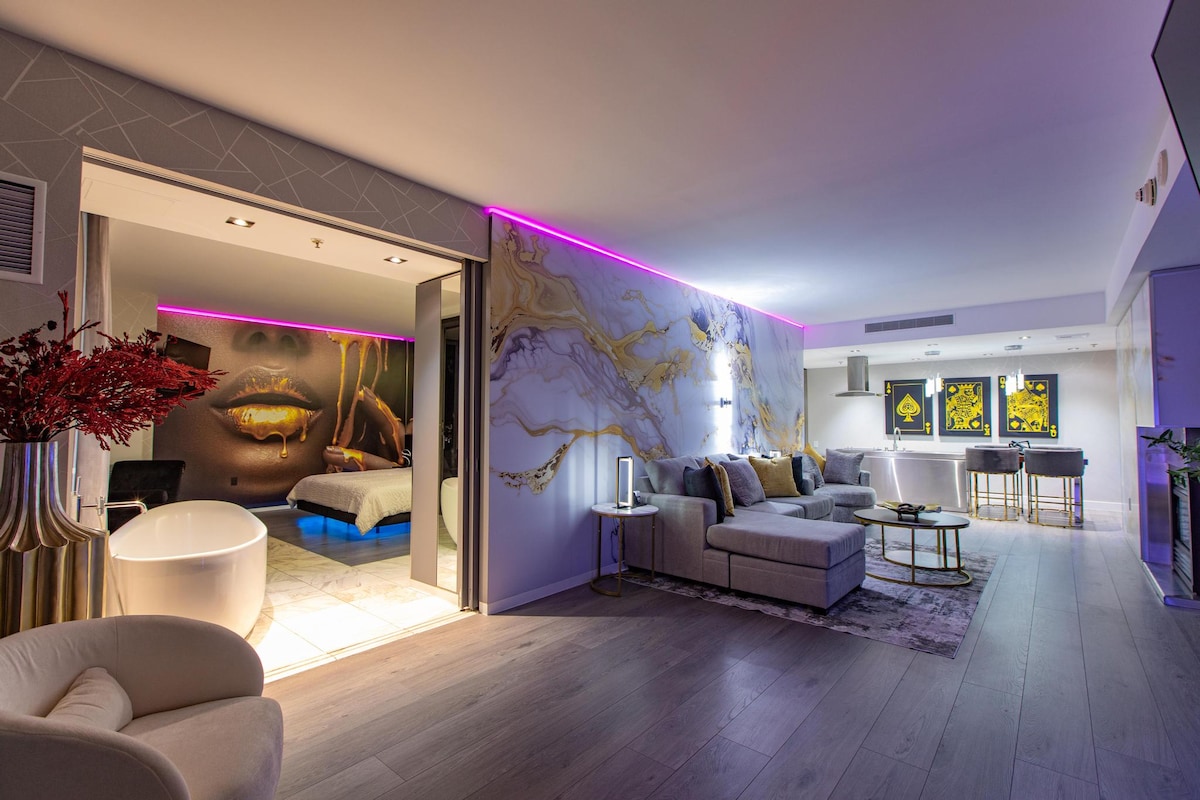 A modern suite is displayed with an open layout, featuring a cozy living area with a sectional sofa and coffee table. A large artwork of lips is showcased on one wall. The bedroom is visible in the distance, along with a stylish bathroom tub illuminated by ambient lighting.