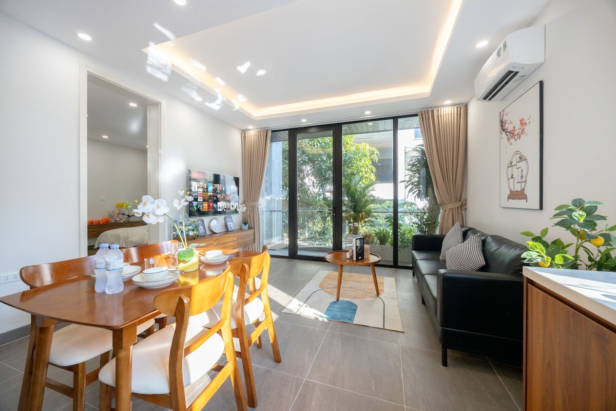 A bright living area features large glass doors that allow natural light to fill the space. A cozy sofa and a wooden dining table set are arranged, complemented by plants and decorative elements. A soft rug lies beneath the coffee table, enhancing the inviting environment.