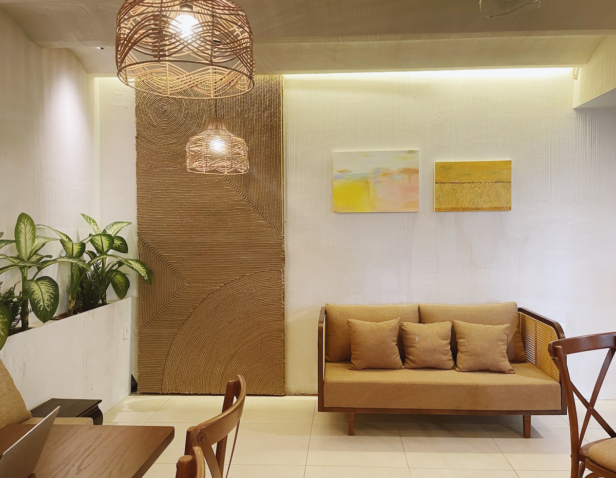 A seating area features a neutral-toned sofa with cushions, positioned against a textured wall. Two art pieces hang above, adding subtle color to the space. Lush green plants are placed in a corner, while warm light from woven pendant fixtures provides a cozy ambiance.