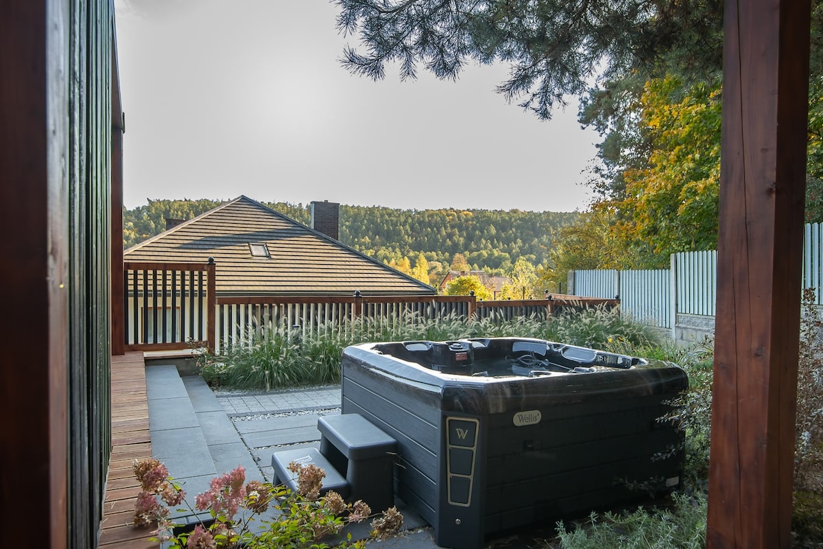 A private hot tub is positioned on a stone patio surrounded by lush greenery. The view reveals a rolling landscape in the background, framed by a wooden fence and colorful foliage. Nearby steps provide access to the hot tub, enhancing the outdoor relaxation space.