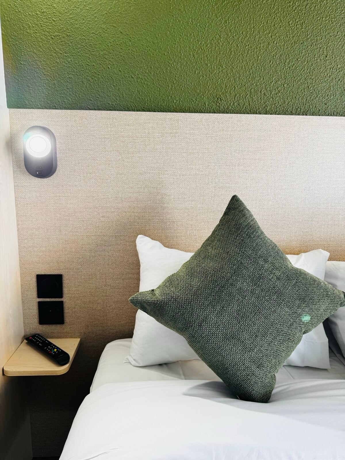 A well-arranged sleeping area features a plush bed with crisp white linens and a large green accent pillow. A small side table holds a remote control, while a light fixture is mounted on the wall above. Textured wall materials add subtle interest.