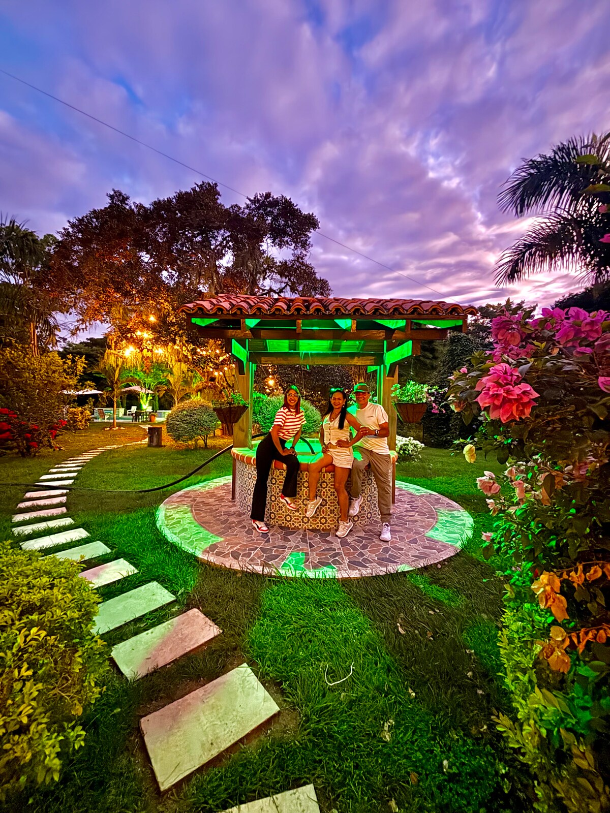 A vibrant garden scene showcases a gazebo surrounded by colorful flowers and lush greenery. Soft lighting highlights the pathway made of stone tiles, leading towards a well-maintained area filled with plants and trees. The sky is adorned with hues of purple and orange as the sun sets.