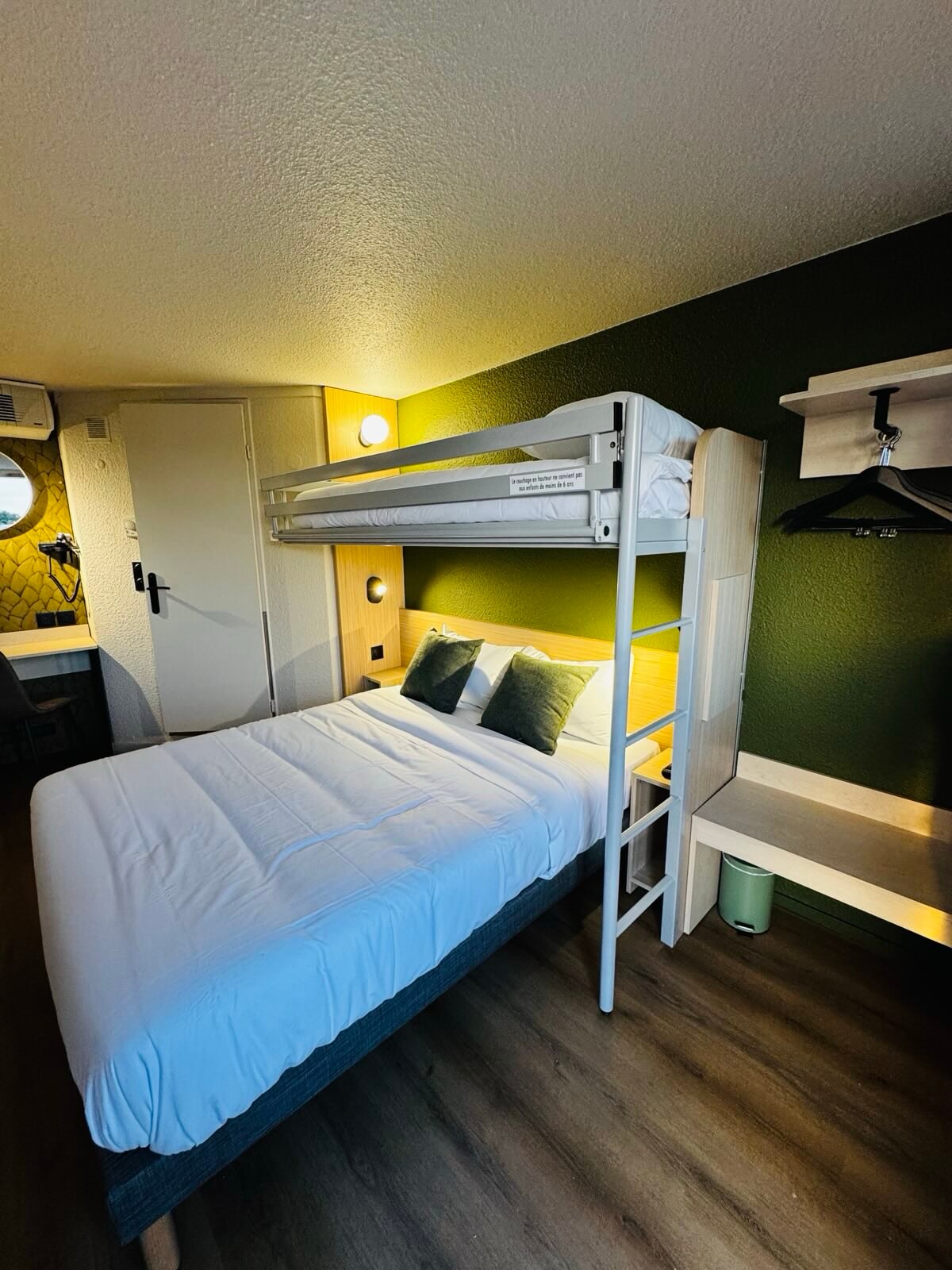 A compact sleeping area features a spacious bed with crisp white linens and two decorative pillows. Above the bed, a lofted bunk is visible. A green accent wall provides contrast, and a minimalist closet space is shown, accompanied by a small bedside lamp.