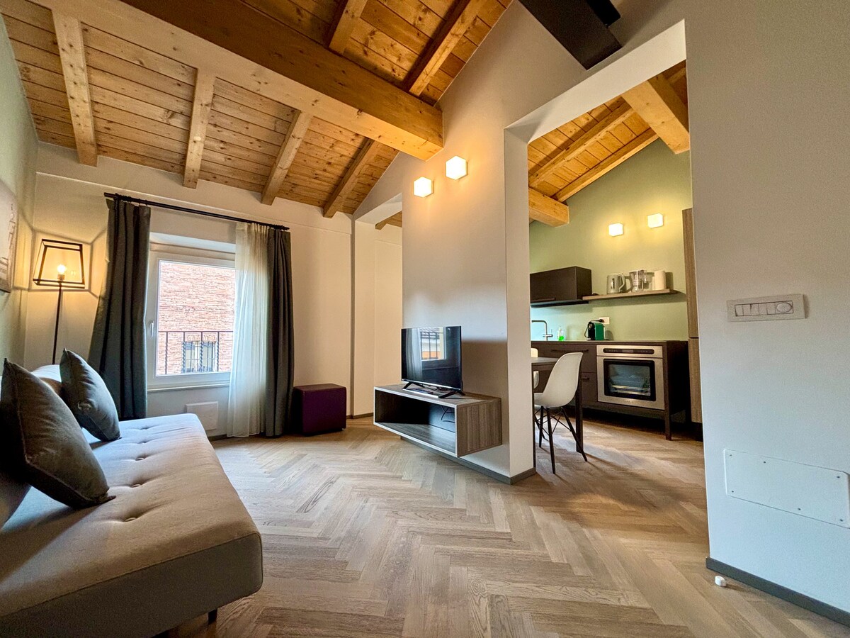 An open space living area features a cozy sofa and wooden ceiling beams that enhance the architecture. A television is positioned in front of the sofa. A fully equipped kitchen is visible in the background, while a large window allows natural light to fill the room.