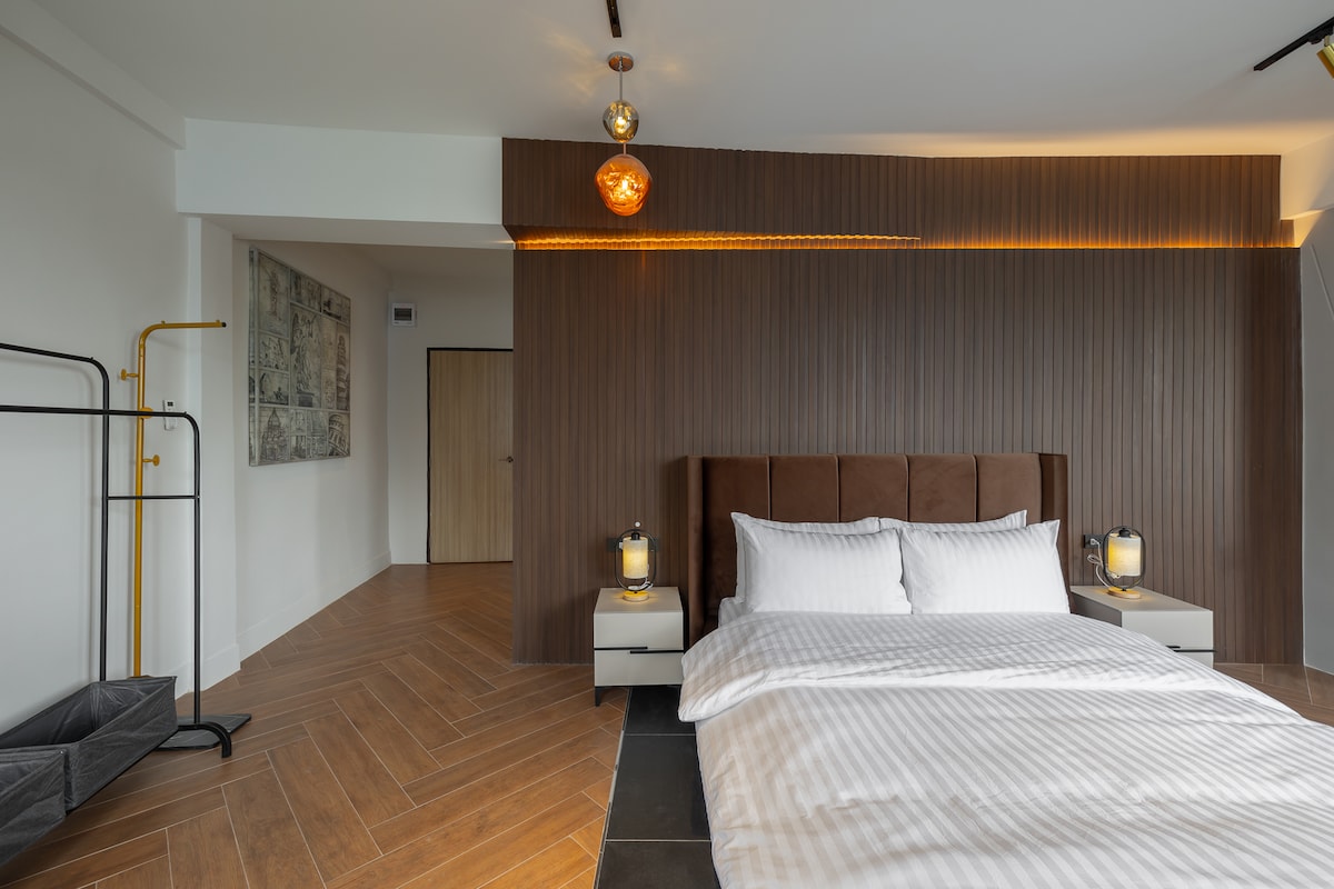 A spacious bedroom is furnished with a large, brown upholstered headboard and striped bedding. Two bedside tables hold lantern-style lamps. A clothing rack stands near the wall, complemented by a modern pendant light above the bed and warm lighting that highlights the wall.