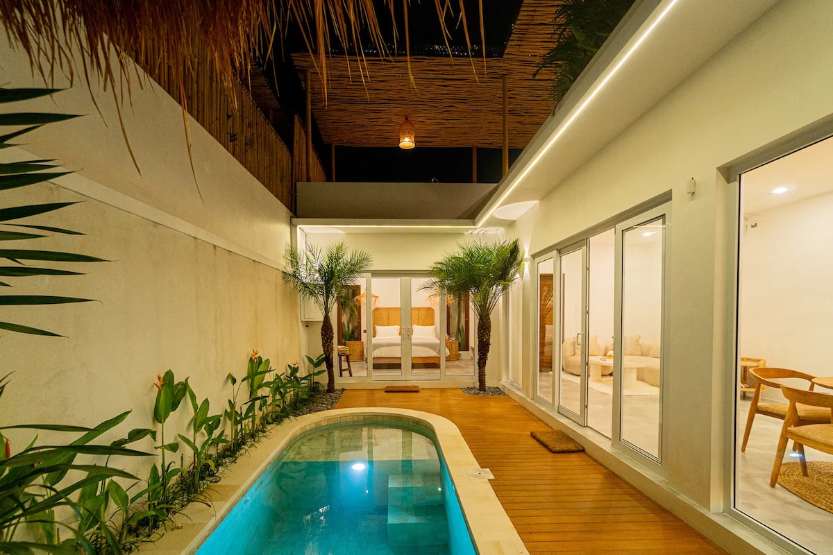 A serene outdoor area features a private pool bordered by wooden decking and tropical plants. Glass doors offer access to a spacious lounge area, illuminated by soft lighting. The natural materials and greenery create a peaceful ambiance, ideal for relaxation.