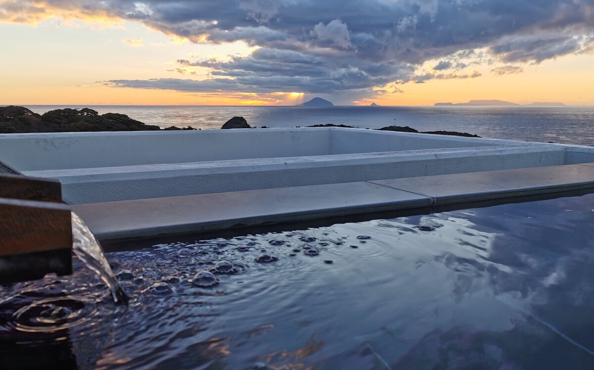 A tranquil outdoor hot spring is positioned along the edge, with water flowing gently from an adjacent spout. The expansive view reveals a vibrant sunset over the ocean, with distant islands visible against the horizon.