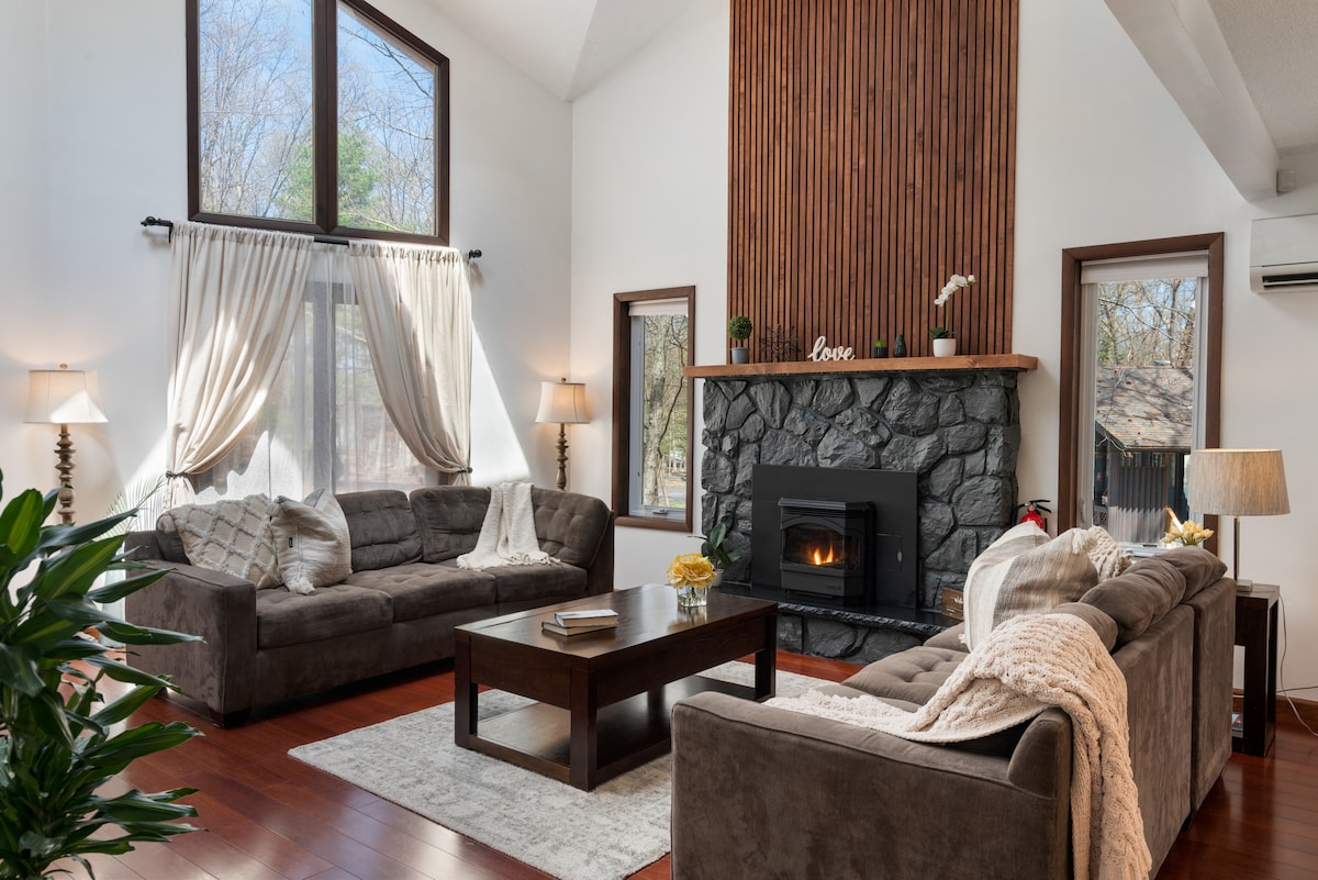 A spacious living room features a stone fireplace adorned with wooden paneling. Two comfortable sofas are arranged around a central coffee table. Large windows allow natural light to fill the space, with soft curtains framing the views. Green plants add a touch of nature.