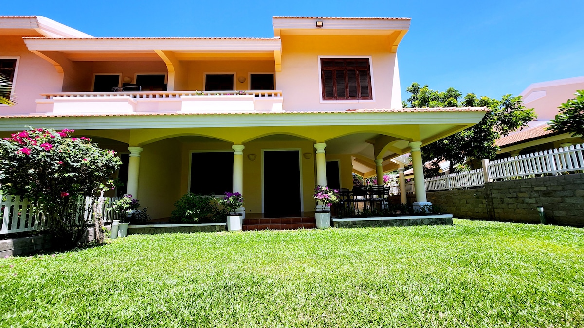 A well-maintained garden surrounds a charming villa with a yellow exterior. The covered patio features several potted plants and a dining area, creating a welcoming outdoor space. The lawn is lush and green, providing a serene view of the villa’s two levels and the clear sky above.