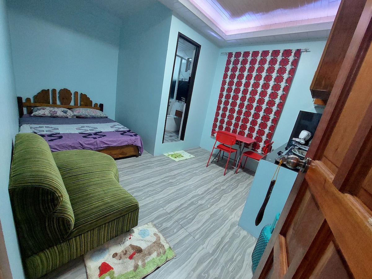 A cozy room features a spacious bed with a patterned blanket and a comfortable green couch. A dining table with two red chairs is set against a wall adorned with colorful patterned curtains. The floor is covered with light-colored tiles, enhancing the room's brightness.