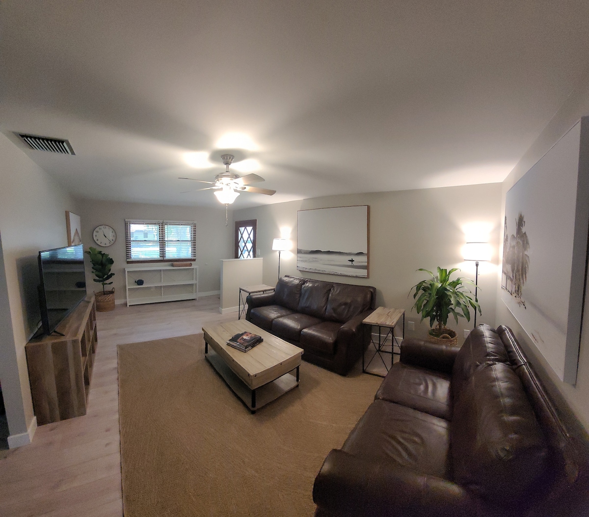 The living area is equipped with two leather sofas facing a wooden coffee table, creating a cozy seating arrangement. A large wall art piece is displayed above the table, while a ceiling fan provides comfort. Natural light enters through the windows, illuminating the space.
