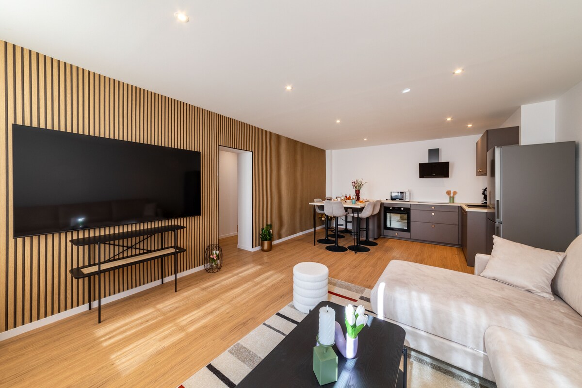 An open-plan living space is presented, featuring a comfortable sofa and a large TV mounted on a wooden accent wall. A modern kitchen area is visible in the background, with a dining table and bar stools. Natural light floods the room through multiple sources.