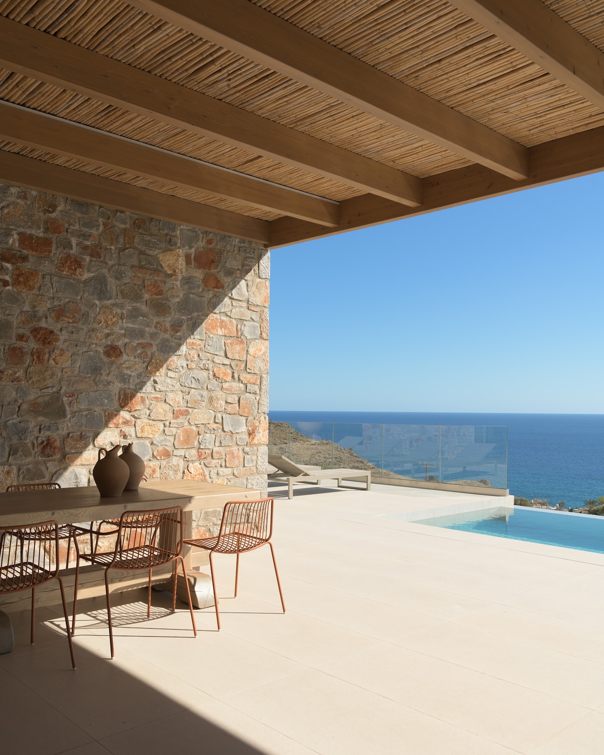 An outdoor dining area features a wooden table with metal chairs, positioned under a covered terrace. The space overlooks a serene sea view, with a sparkling pool visible nearby. Natural stone walls add texture to the setting, while sunlight creates a bright ambiance.