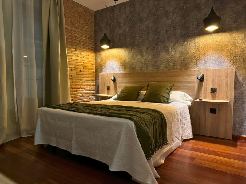 A double room features a spacious bed dressed with a white coverlet and two green accent pillows. Wood accents are present in the headboard and nightstands, which are lit by wall-mounted lamps. Natural light filters through sheer curtains, illuminating the warm wooden floors.