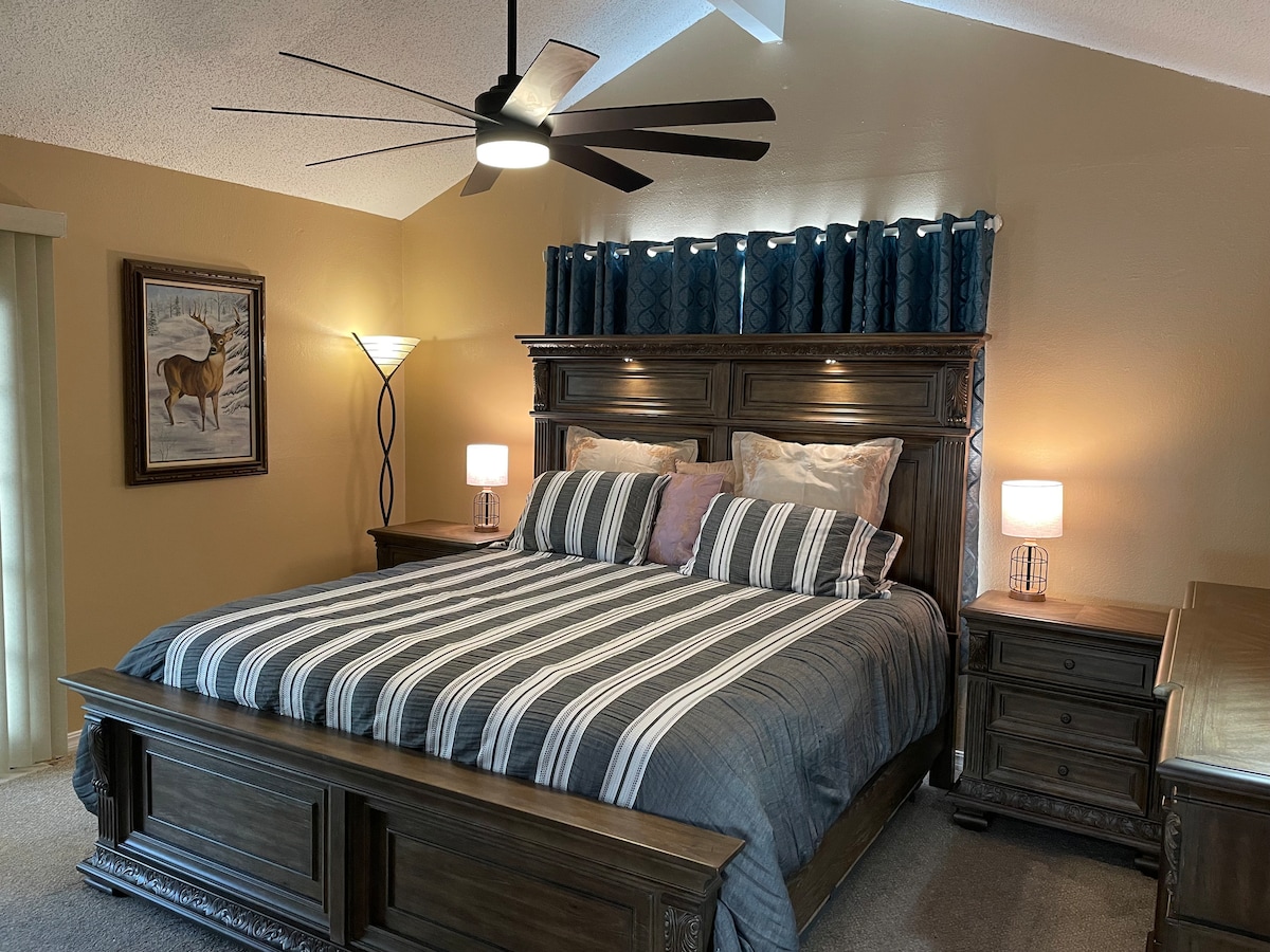 The master bedroom features a spacious bed with a striped duvet and decorative pillows. Flanking bedside lamps provide soft lighting, while a ceiling fan adds comfort. A large piece of artwork depicting a deer is hung on the wall, complemented by decorative curtains above the headboard.