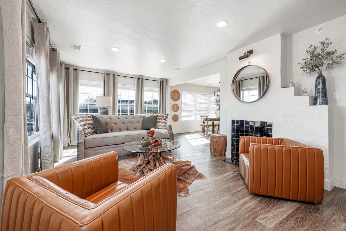 A light-filled living area is defined by large windows adorned with sheer drapes. A comfortable gray sofa is complemented by two warm-toned leather chairs. A glass coffee table, set atop a textured rug, is centrally placed on the wood flooring, providing a cozy atmosphere.