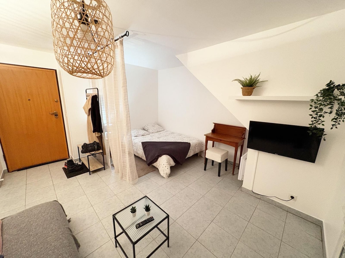 A studio space features a cozy sleeping area with a spacious bed covered in neutral bedding. A small wooden desk is positioned next to a television mounted on the wall. A stylish pendant light hangs from the ceiling, and a sheer curtain divides the space. Neutral tile flooring enhances the bright ambiance.