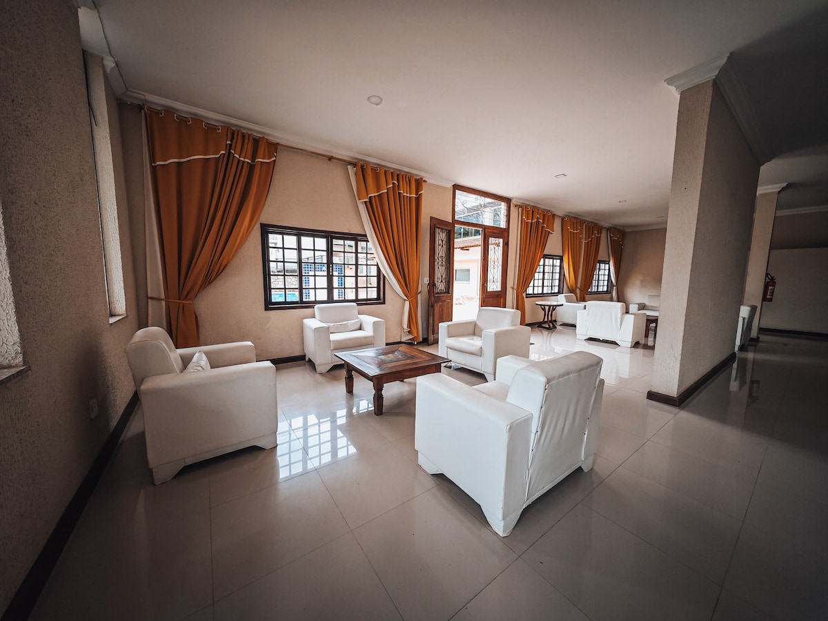 A spacious common area is furnished with white chairs arranged around a wooden coffee table. Large windows allow natural light to fill the room, complemented by light-colored curtains hanging from the walls.