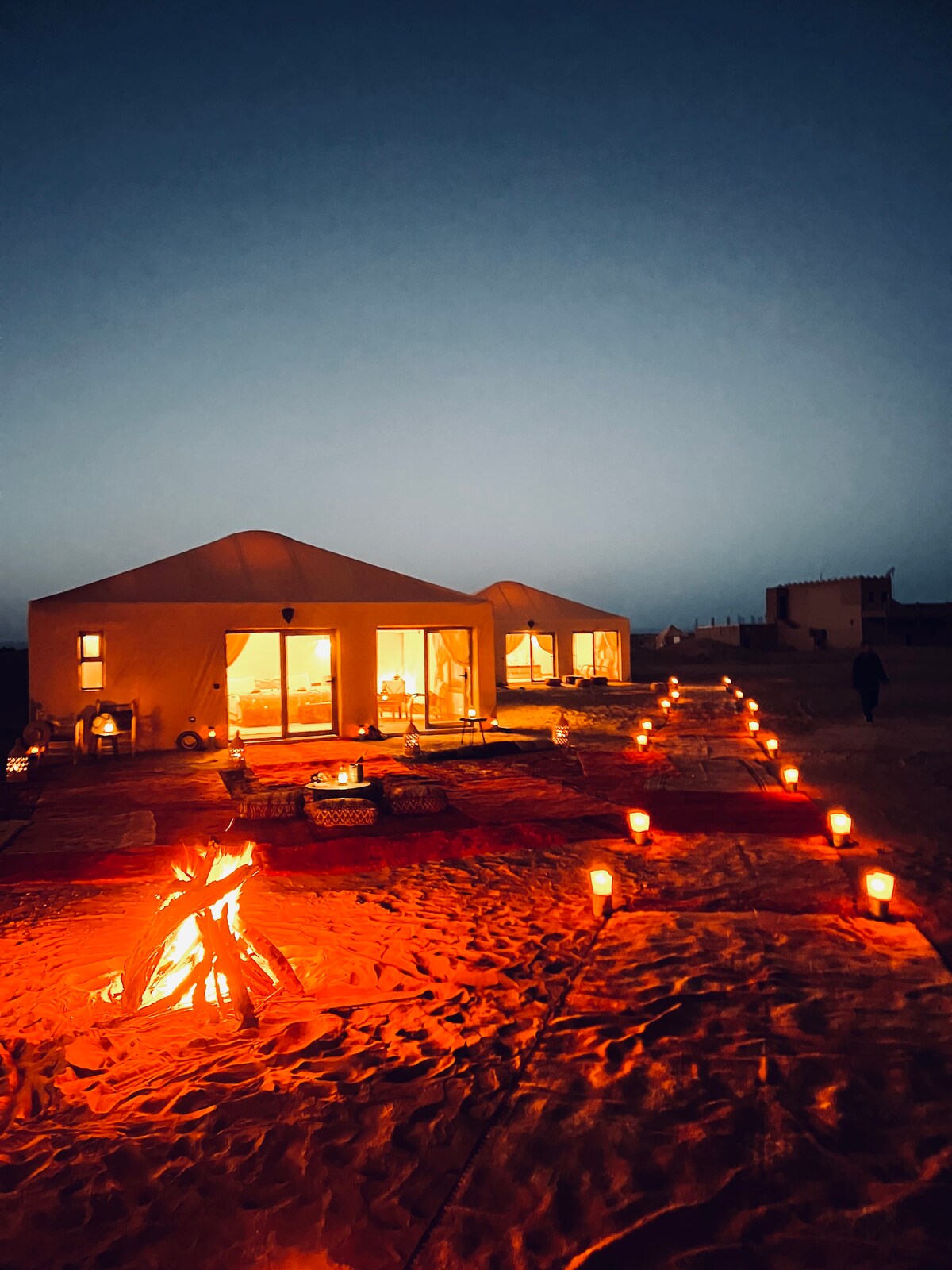 Two luxurious tents are softly illuminated at dusk, with warm light spilling from their large windows. A fire pit is surrounded by seating, and lanterns line a sandy path leading to the tents, creating a serene and inviting atmosphere in the desert.