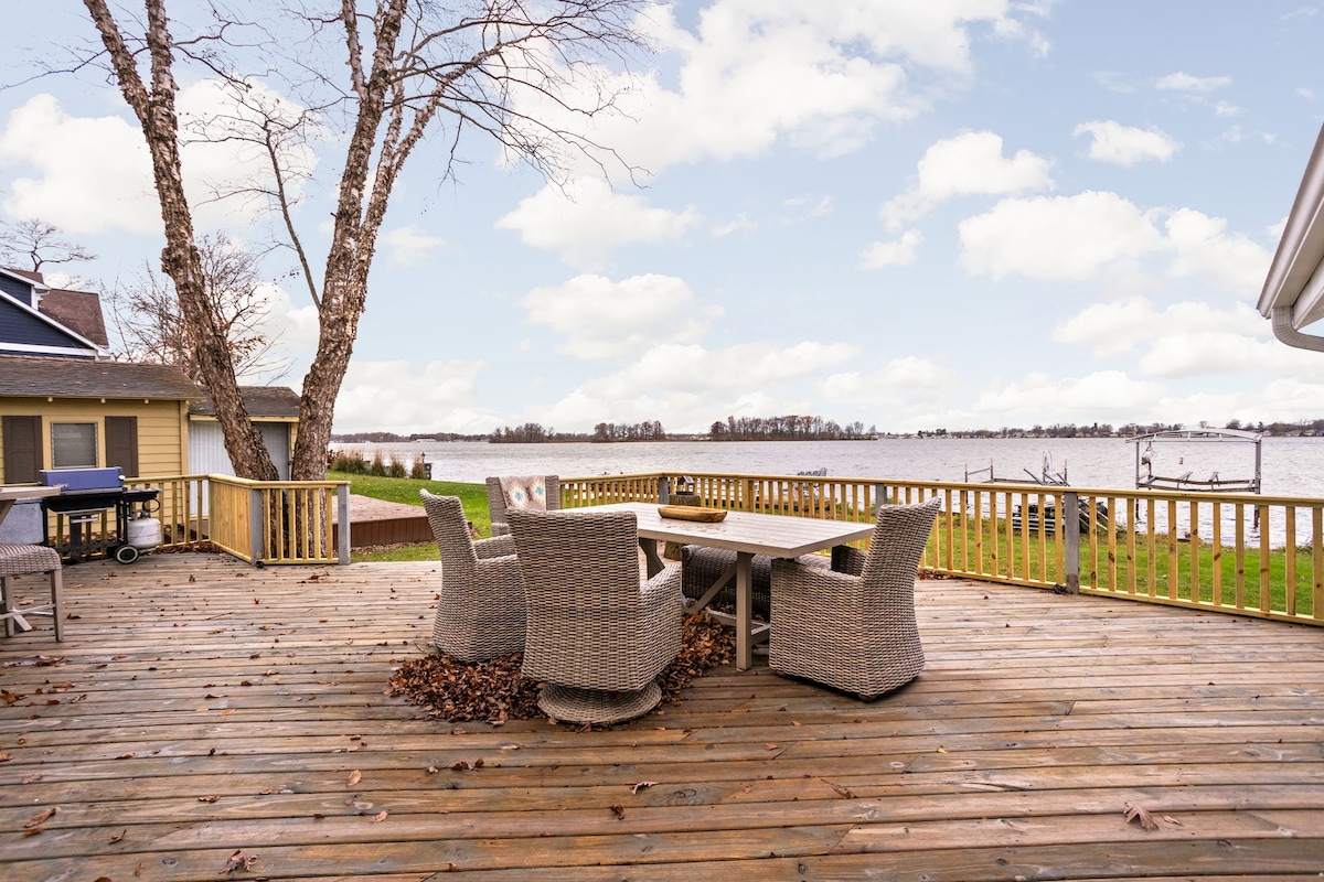 An expansive wooden deck is featured, surrounded by nature, with a large table and six wicker chairs arranged for outdoor dining. A grill is positioned nearby, and the serene lake is visible in the background, creating a peaceful setting.