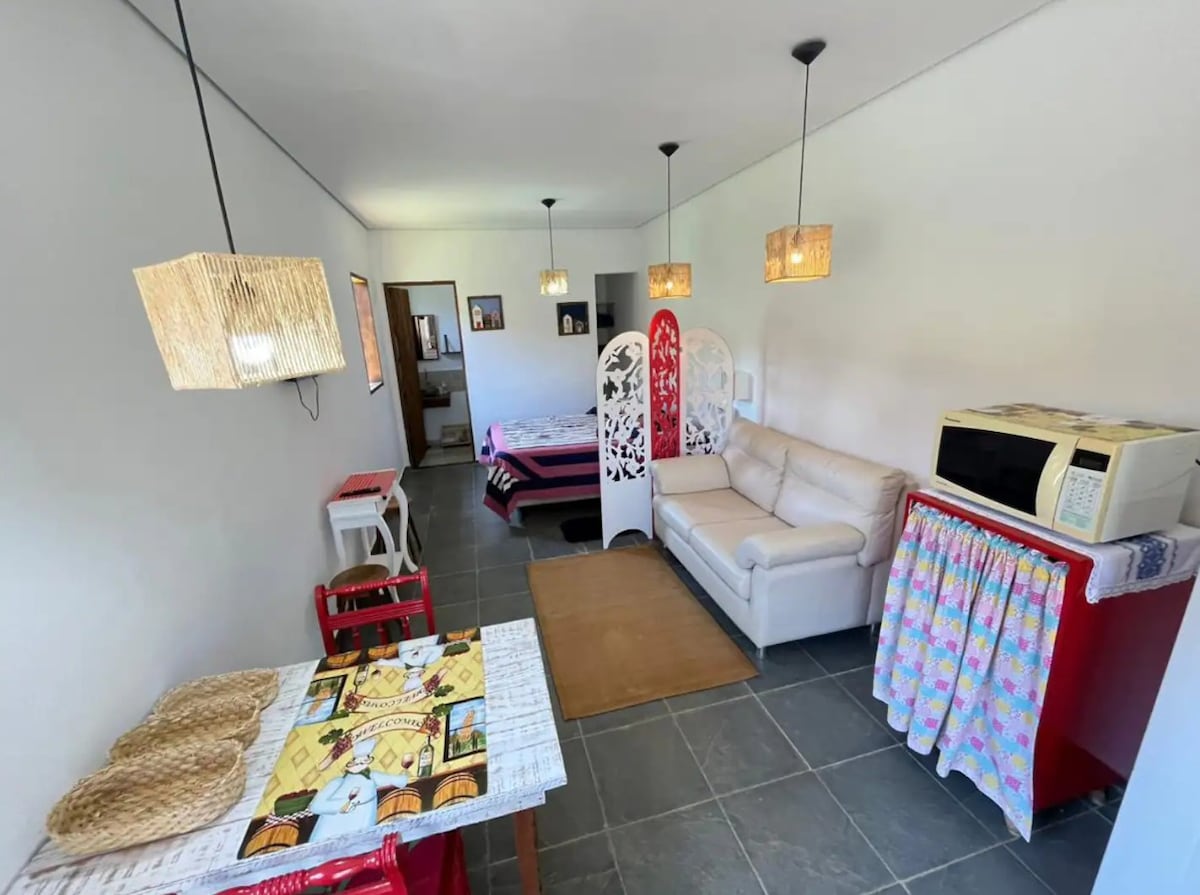 An open living space features a cozy sofa and a dining area with a table and red chairs. Decorative lamps hang from the ceiling, adding warmth. A microwave and colorful cloth cover are positioned on a small cabinet, with a doorway leading to the bedroom visible in the background.