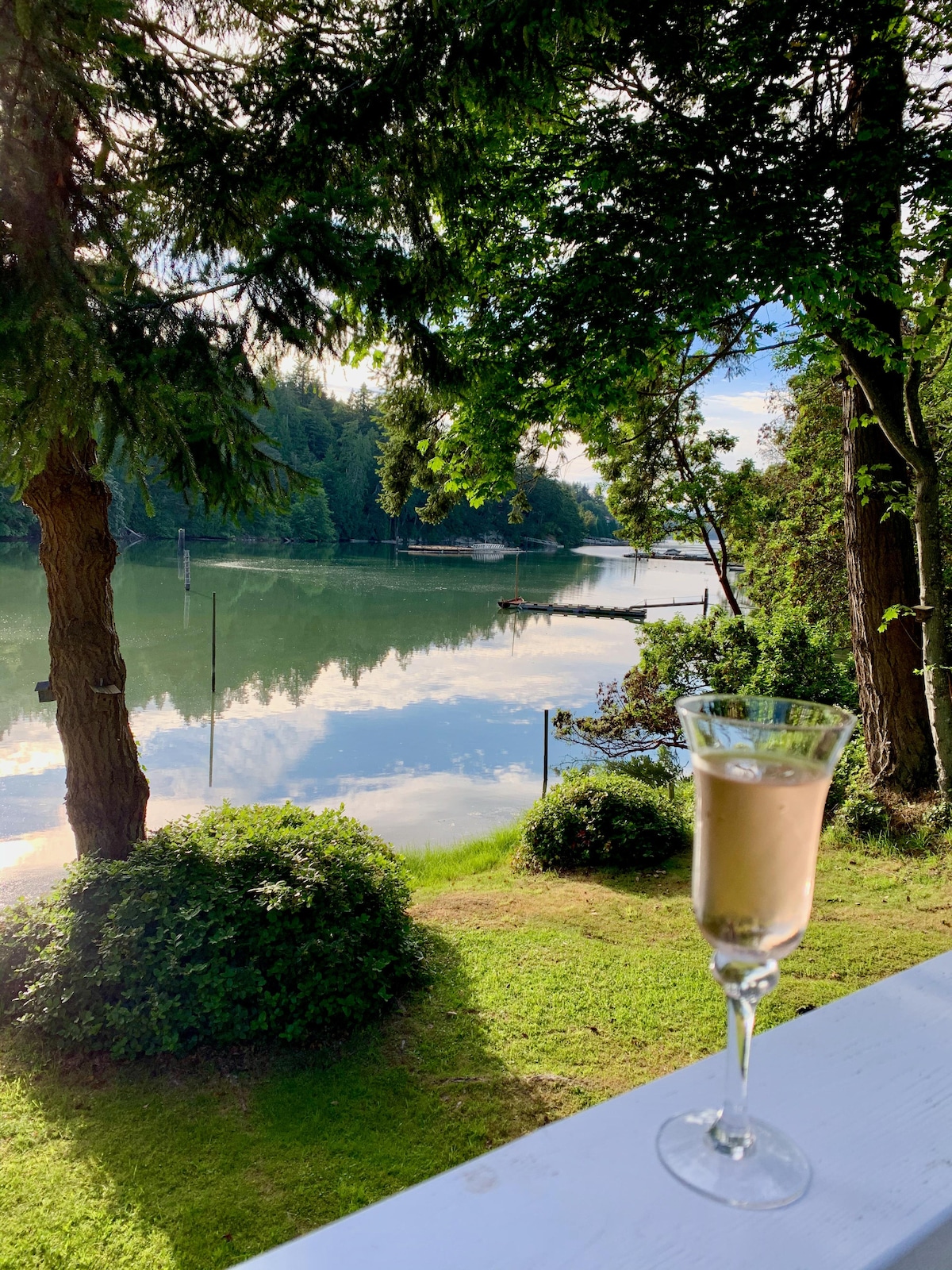 A serene view of the calm water reflecting the trees and sky is presented. In the foreground, a glass filled with a light beverage stands on a railing. Lush greenery frames the scene, enhancing the peaceful ambiance.