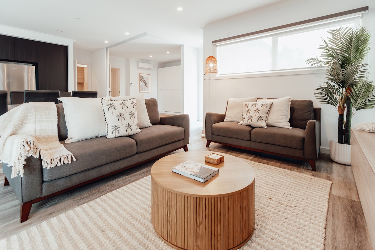 A modern living area features two comfortable sofas adorned with decorative pillows, positioned around a round wooden coffee table. A textured area rug complements the space, while large windows allow natural light to illuminate the room. A tall potted plant adds a touch of greenery.