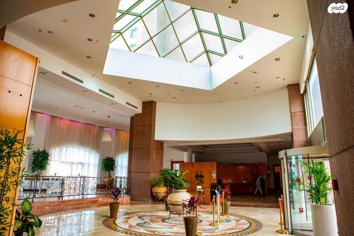 An expansive lobby area is illuminated by natural light streaming through a sky dome. A decorative tile floor features a circular pattern at the center. Lush plants are arranged throughout the space, complemented by elegant furniture along the walls.